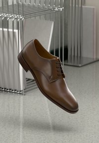 Scarpa di pelle marrone con lacci, punta arrotondata, superficie liscia, dettagli di cucitura sottili e suola in gomma marrone medio, sospesa sopra un pavimento.