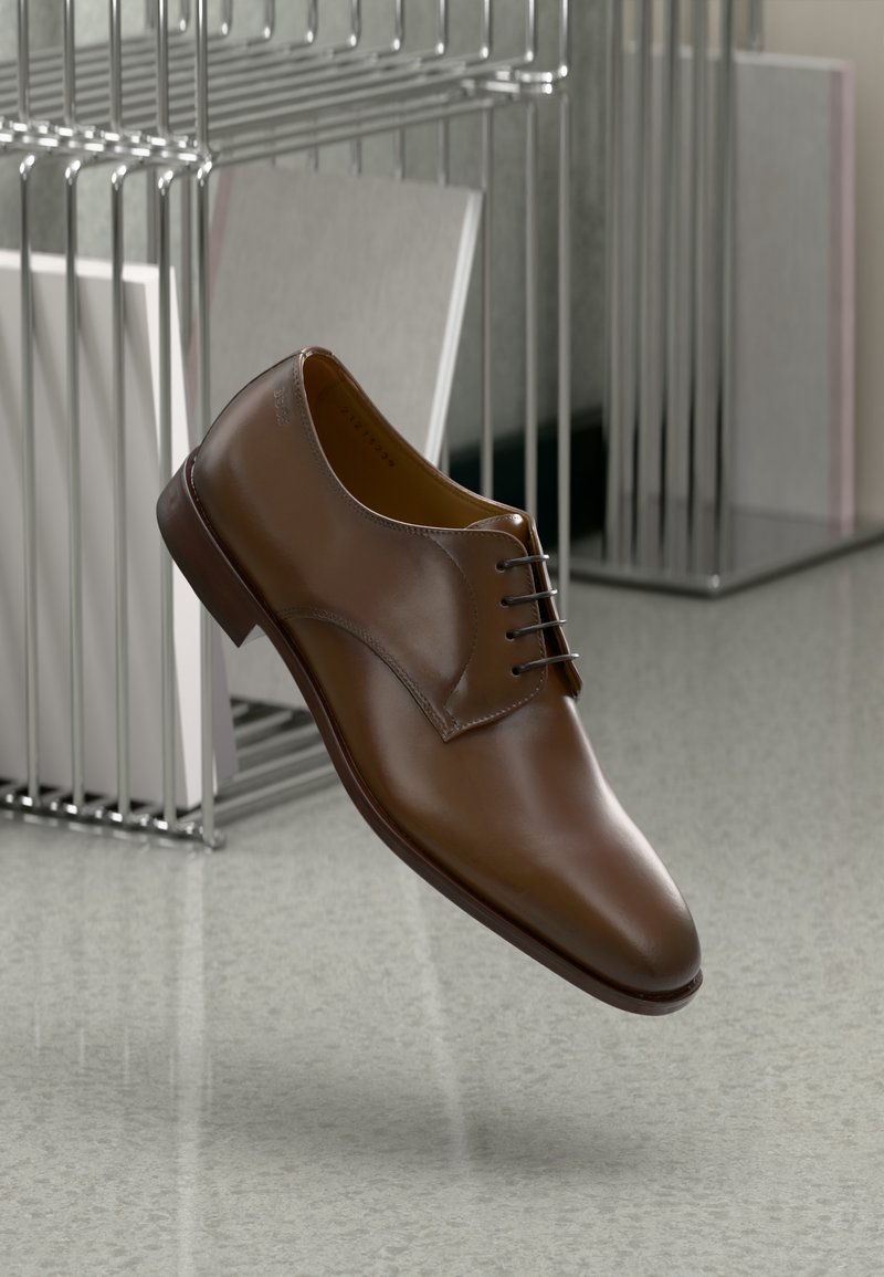Scarpa di pelle marrone con lacci, punta arrotondata, superficie liscia, dettagli di cucitura sottili e suola in gomma marrone medio, sospesa sopra un pavimento.