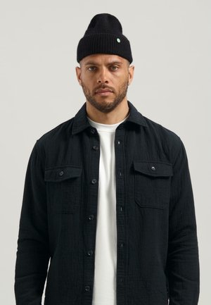 Zwarte overhemd met knopen en twee borstzakken, textuurstof. Witte ronde hals t-shirt eronder. Zwarte geribbelde beanie met een groene accent.