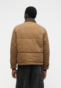 Jachetă din material padding de culoare brună, cu guler din catifea. Are un design matlasat și capse metalice la manșete. Textură netedă și croială lejeră.