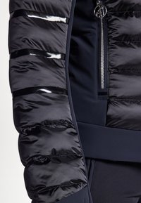 Tumesin puffer-jakk navy värvi, läikivate mustade detailidega. Omab tikandiga osi varrukatel, siledat tekstuuri ja lukuga taskut.