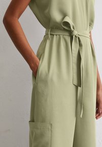 Intramontabile tuta verde chiaro con vita con cintura, maniche corte, tasche laterali e una texture di tessuto liscia, che evidenzia una vestibilità rilassata.