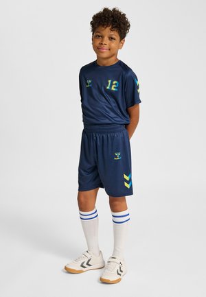 Junge steht mit hinter dem Rücken verschränkten Händen, trägt marineblaue Sportkleidung mit der Nummer 12, weiße Socken mit blauen Streifen und weiße Turnschuhe.