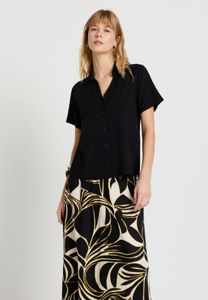 Donna che indossa una camicia nera a maniche corte con bottoni e una gonna bianca e nera con un grande motivo a foglie.