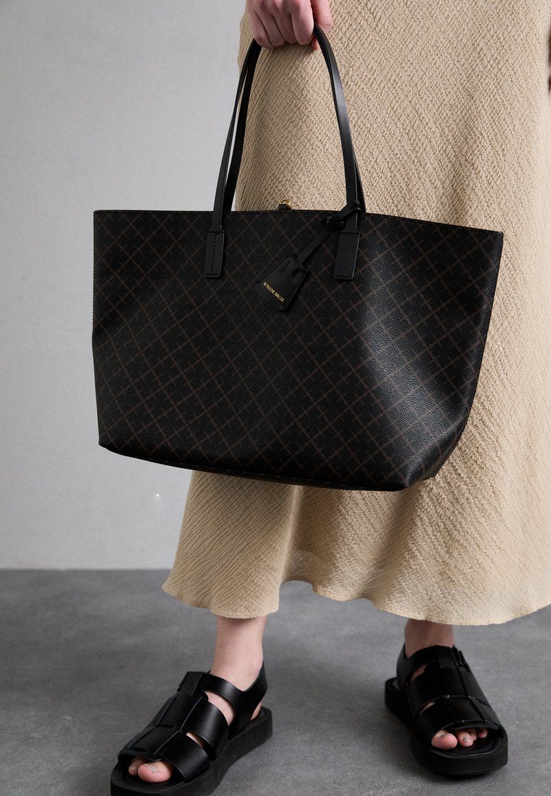Black tote bag with a geometric pattern, featuring two black handles and a small tag. Worn with a textured beige skirt and black sandals.