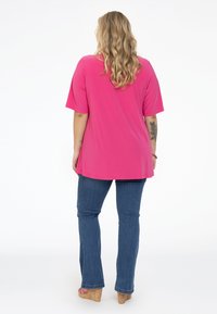 Rosa Kurzarmoberteil mit lässigem Schnitt, kombiniert mit dunkelblauen Schlagjeans und Kork-Plateau-Pumps. Die Haare sind wellig und schulterlang.