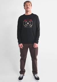 Chemise noire à manches longues avec un motif graphique comprenant des accents rouges, beiges et noirs. Associée à un pantalon marron et des baskets noires.