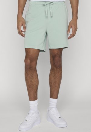 Mężczyzna noszący jasnzie zielone spodnie z sznurkiem, białe skarpetki i białe trampki, stojący na tle jednolitego tła.