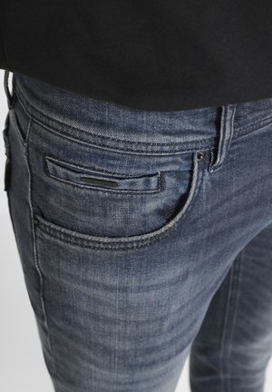 Jeans de mezclilla azul oscuro con una textura desgastada, que cuentan con un bolsillo delantero y bolsillos traseros, acentuados con costuras y herrajes en negro.