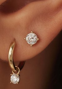 Les boucles d'oreilles créoles en or présentent une pierre précieuse ronde et transparente en haut, avec une plus petite suspendue en dessous, mettant en valeur une finition polie et un éclat brillant.