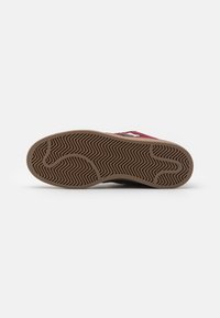 Sneaker na gumovej podrážke s hnedou textúrovanou spodnou časťou a červeným zvrškom s bielym brandingom. Dizajn obsahuje vzor zubov v tvare zigzag.