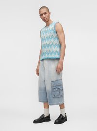 Homme debout portant un haut tricoté sans manches bleu et blanc à motifs, un short large en denim clair avec de grandes poches, des chaussettes blanches et des chaussures noires.