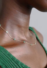 Zelta ķēdes kaklarota ar apaļiem zaļiem akmeņiem, kas izkārtoti maiņus. Delikāts dizains guļ uz ādas, papildināts ar zaļu auduma fonu.