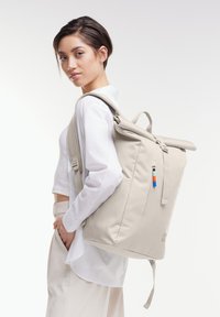 Sac à dos beige à rabat avec des bretelles réglables, au design épuré, en tissu texturé, avec une poche à zip sur le côté et des accents de fermeture éclair contrastants.