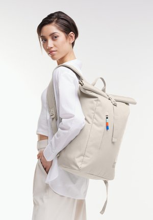 Sac à dos beige à rabat, avec des bretelles ajustables, une poche zippée à l'avant et une étiquette d'accent colorée, fabriqué en tissu résistant.