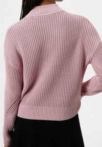 Różowy sweter robiony na drutach o teksturowanym, luźnym splocie, z prążkowanym kołnierzem i mankietami oraz krótkim krojem. Tył ma czysty design bez ozdób.