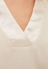 Top beige con escote en V, que presenta un cuello de satén en contraste y una tela texturizada, mostrando un sutil brillo y un acabado limpio en las costuras.