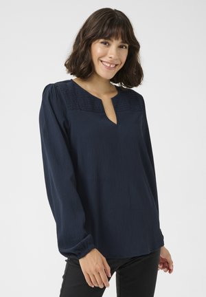 Blouse bleu marine à manches longues avec un col en découpe, panneau supérieur texturé et poignets froncés. Tissu lisse avec une coupe décontractée.