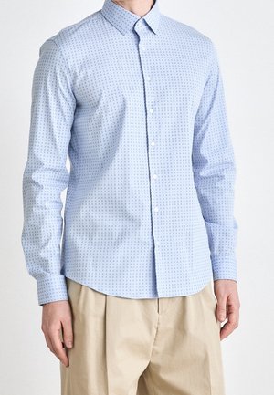 Mann trägt hellblaues, langärmeliges Hemd mit kleinen, dunklen Anker-Mustern und beigen, lockeren Hosen, steht vor einfarbigem Hintergrund.