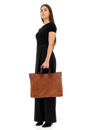 Brown leather tote bag with decorative stitching, dual round handles, and a spacious design, held by a person in black attire.