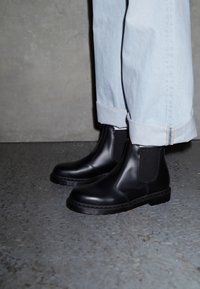 Las botas Chelsea negras se exhiben junto a unos jeans azules claros y doblados. El fondo es una superficie gris texturizada.