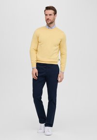Męski jas yellow sweter, okrągły dekolt, dziana tekstura, połączony z ciemnoniebieskimi spodniami i białymi sneakersami. Minimalistyczny design, brak wzorów.