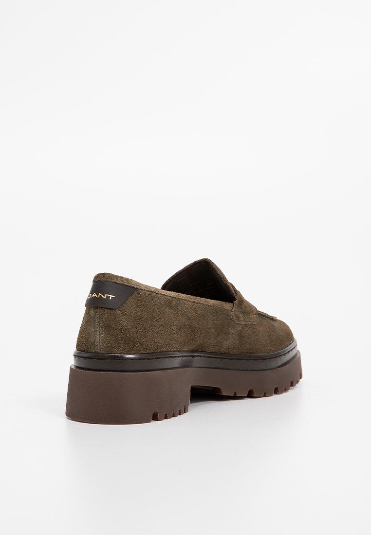 Mocassino in suede marrone con suola spessa e linguetta nera sul tallone che presenta il logo dorato del marchio, mostrato dal lato posteriore su sfondo bianco.