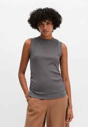 Femme aux cheveux bouclés portant un top gris sans manches et un pantalon beige, debout avec une main dans la poche sur un fond uni.