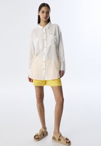 Weißes Langarmhemd mit Knopfschluss und zwei Brusttaschen, kombiniert mit ausgefransten gelben Shorts. Das Modell trägt beigefarbene Sandalen.