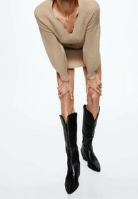 Schwarze Lederstiefel bis zum Knie mit spitz zulaufender Zehe, angularen Ausschnitt-Design am oberen Teil und einem stabilen, erhöhten Absatz. Die Textur wirkt glatt.