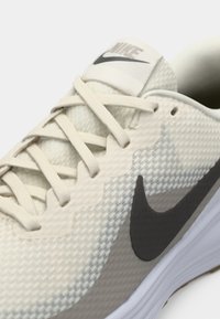 Gros plan sur une chaussure de course Nike beige avec lacets blancs, tissu texturé, et le logo Nike noir sur le côté.