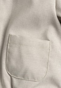 Nabij een beige stof, waarbij een rechthoekige zak met zichtbare stiksels is te zien. De textuur is glad met een subtiel weefpatroon.