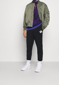 Olivgrön bomberjacka med ribbade ärmslut, svarta träningsbyxor med blå accenter och vita sneakers. Jackan har en sid ficka och logotyp.