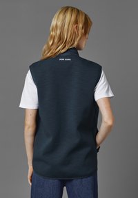 Marinblå ärmlös väst med en mjuk textur, som har en hög krage och en vit "PEPE JEANS"-logotyp på ryggen, bärs över en vit t-shirt.