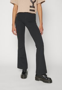 Svarta utsvängda byxor i ett stretchigt material, tillsammans med en beige oversize-skjorta med djärva svarta bokstäver. Svarta kraftiga kängor fulländar outfiten.