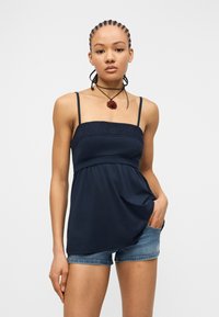 Νέα γυναίκα με πλεγμένα μαλλιά, φοράει μπλε μπλουζάκι καμησόλ και τζιν σορτς, στέκεται με το ένα χέρι στην τσέπη μπροστά σε απλό φόντο.