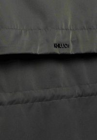 Detail tmavě olivově zelené péřové bundy s horizontálním prošitím a malým černým vyšitým logem "KHUJO" poblíž středního švu.