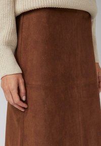 Gros plan sur des mains reposant à côté d'une jupe en daim marron portée avec un pull en laine crème.