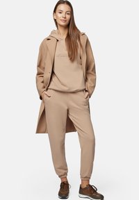 Jas_beżowy dres z kapturem i dresami z elastycznymi mankietami, nałożony pod długim beżowym płaszczem. Brązowe trampki dopełniają strój.
