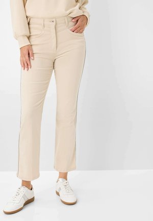 Personne portant un pantalon droit beige, un sweat crème et des baskets blanches, debout devant un fond clair uni.