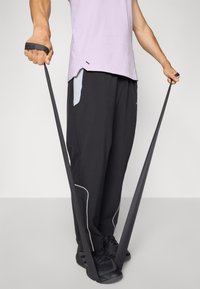 Zwarte sportbroek met witte accenten en zijzakken, gecombineerd met een lichtpaarse shirt. Een weerstandsband wordt in beide handen vastgehouden.