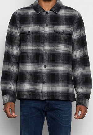 Chemise à carreaux noire et grise avec boutons et poches poitrine portée sur un jean bleu foncé par une personne.