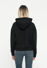 Svart hettegenser med en avslappet passform, som har en stor hette, ribbede mansjetter og kant, kombinert med tettsittende blå denimjeans.
