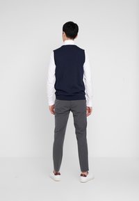 Marineblaues ärmelloses Weste über einem weißen Hemd, kombiniert mit grauen, zulaufenden Hosen und weißen Turnschuhen. Glatter Stoff, schlichtes Design, keine Muster.