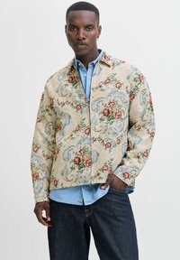 Bloemmotief jas in lichtbeige stof, met rode en blauwe bloemen, gecombineerd met een lichtblauwe spijkerblouse en donkere jeans.