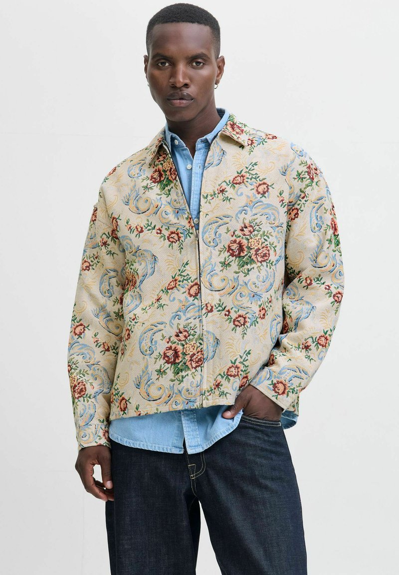 Bloemmotief jas in lichtbeige stof, met rode en blauwe bloemen, gecombineerd met een lichtblauwe spijkerblouse en donkere jeans.