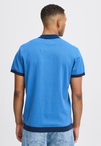 Kék, rövid ujjú pólóing, tengerészkék bordázott gallérral és mandzsettával, sima tapintású és egyenes aljú. Nincsenek látható logók vagy minták.