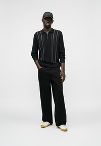 Μαύρο φούτερ με φερμουάρ και κάθετες γκρι ρίγες, συνδυασμένο με φαρδιά μαύρα παντελόνια και λευκά αθλητικά παπούτσια με μπεζ σόλες, ολοκληρωμένο με ένα μαύρο καπέλο.