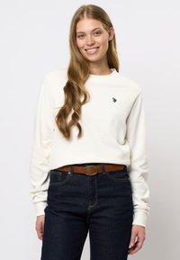 Jonge vrouw met lang golvend haar, gekleed in een wit shirt met lange mouwen met een klein logo en donkerblauwe jeans met een bruine riem, glimlachend.