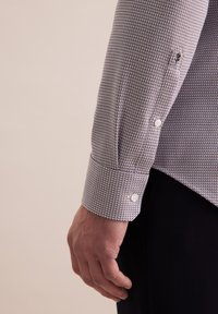 Karierter Hemdenärmel in Weiß und Dunkelblau, mit silbernen Knöpfen und einer subtilen Textur. Nahaufnahme einer Hand, die neben dem Ärmel ruht.